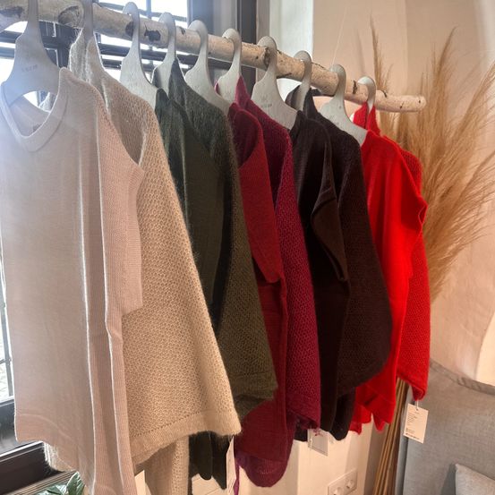 Eine Reihe von Pullovern in verschiedenen Farben, darunter weiß, beige, grün, rot und braun, hängt an einem Holzständer an einer weißen Wand.