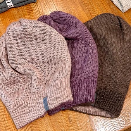 Drei gestrickte Mützen liegen auf einem Holztisch. Die Farben sind hellrosa, lila und braun. Ein grauer Anhänger mit 'KATTIO' ist an der rosa Mütze sichtbar.