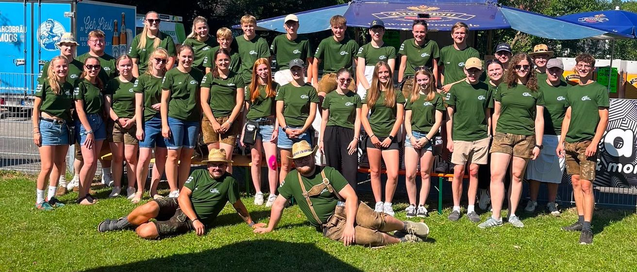 Eine Gruppe von Menschen in grünen Shirts und Hüten posiert für ein Foto in einem Park mit Bäumen und Schirmen im Hintergrund.