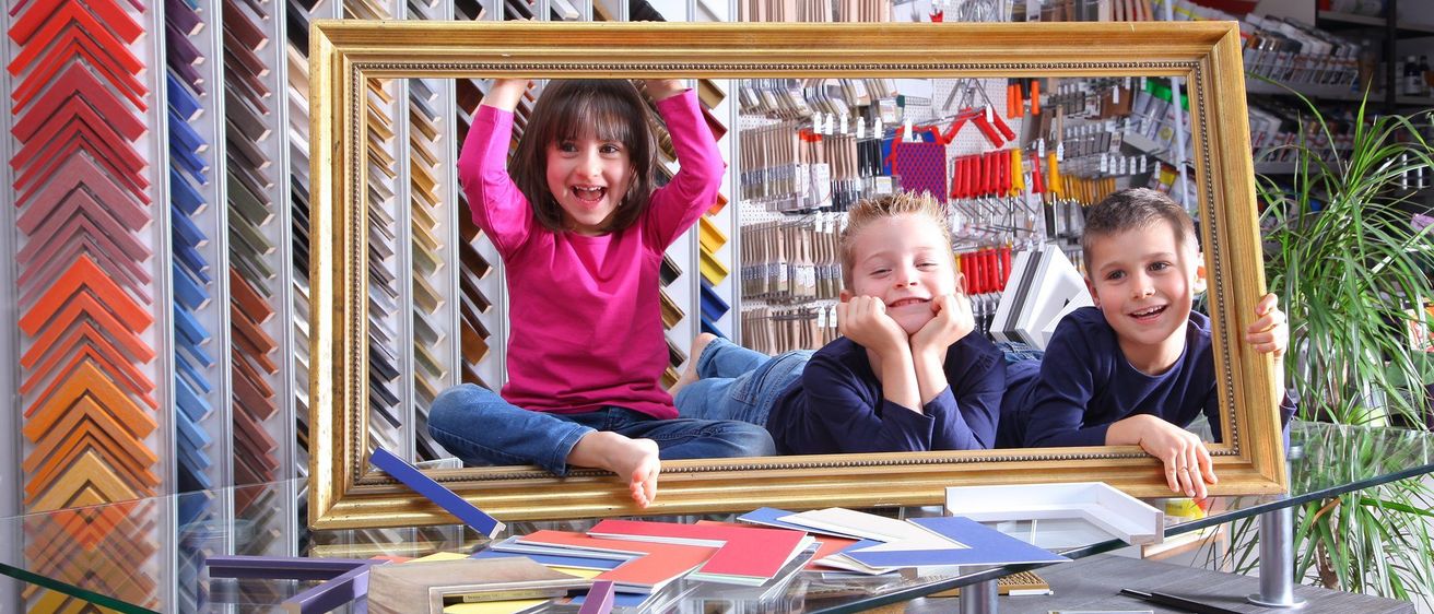Drei Kinder posieren in einem goldenen Bilderrahmen in einem Geschäft. Sie lächeln und scheinen für ein Foto zu posieren. Der Tisch ist mit verschiedenen Rahmen gefüllt.