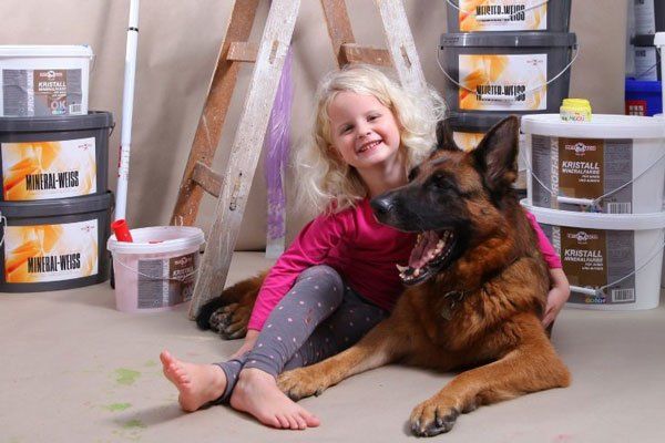 Ein kleines Mädchen mit blonden Haaren sitzt auf dem Boden neben einem deutschen Schäferhund, umgeben von Farbbechern und einer Leiter.