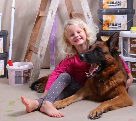 Ein kleines Mädchen mit blonden Haaren sitzt auf dem Boden neben einem deutschen Schäferhund, umgeben von Farbbechern und einer Leiter.