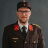 Ein Mann in Uniform mit Brille und Hut lächelt für ein Foto.
