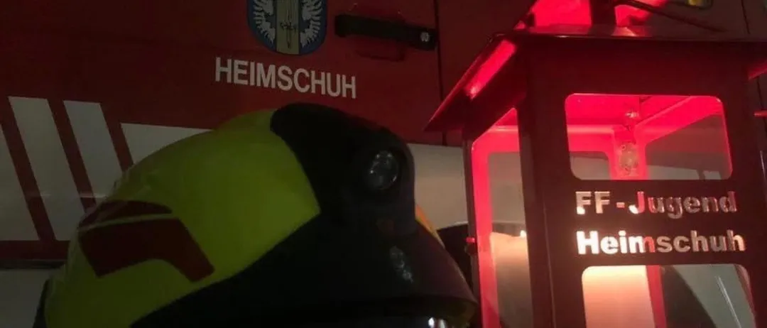 Ein Feuerwehrhelm und eine rote Laterne mit den Worten FF-Ju Heimschuch stehen neben einem Feuerwehrwagen. Der Wagen hat die Aufschrift Freiwillige Feuerwehr oben.