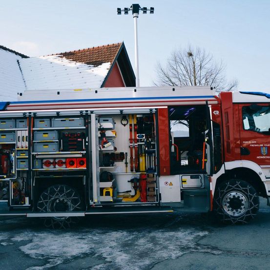 Ein roter Feuerwehrwagen mit Ketten an den Rädern steht vor einem Gebäude mit verschneitem Dach.