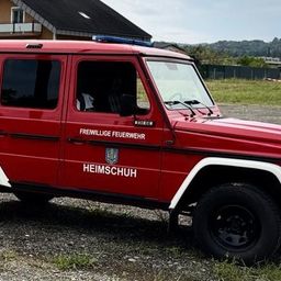 Ein rotes Feuerwehrfahrzeug mit 'KDO' und 'FREIWILLIGER FEUERWEHR' Markierungen ist auf Kies geparkt. Dahinter befindet sich ein Rasenbereich mit Häusern und Bäumen.