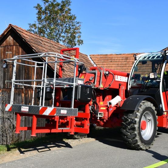 Ein roter Traktor mit einem großen roten Korb auf dem Rücken steht vor einem Holzhaus mit braunem Dach.