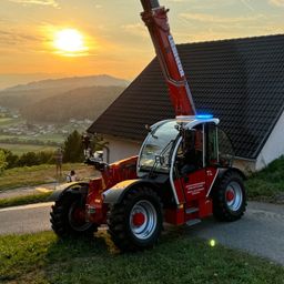 Ein roter Traktor mit einem Kran steht auf einem Rasen neben einem Haus. Der Traktor hat oben ein blaues Licht. Eine Person sitzt drinnen. Die Sonne geht in der Ferne unter.