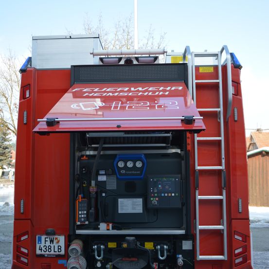 Rückansicht eines roten Feuerwehrfahrzeuges mit offener Luke, auf der der Schriftzug 'FEUERWEHR HEIMSCHUH' steht. Eine Leiter ist an der Seite befestigt. Das Nummernschild lautet 'FW 438 LB'.
