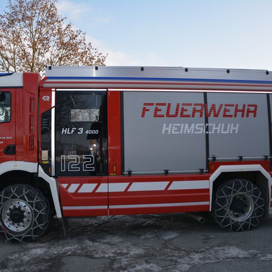 Ein roter und weißer Feuerwehrwagen mit der Nummer 122 ist in einem verschneiten Bereich geparkt. Der Wagen hat eine graue Tür mit den Worten 'FEUERWEHR HEIMSCHUH'.