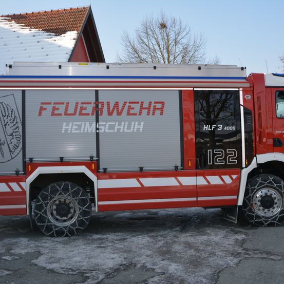 Ein roter Feuerwehrwagen mit 'FEUERWEHR HEIMSCHUH' und 'HLF 3 4000 122' steht vor einem Haus mit Schnee auf dem Dach.
