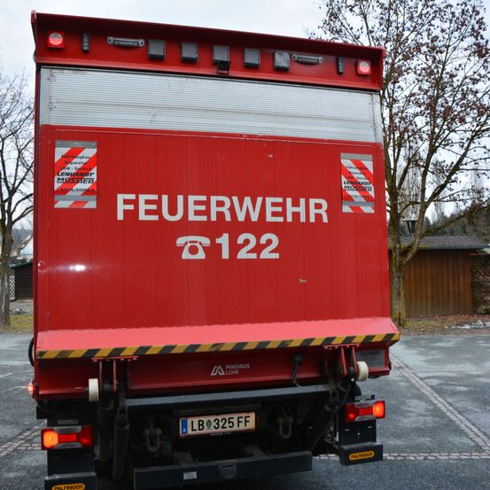 Die Rückseite eines roten Feuerwehrfahrzeuges mit den Worten 'FEUERWEHR 122' und einem Kennzeichen 'LB 325 FF'. Es hat rote Notleuchten und einen reflektierenden Warnstreifen.