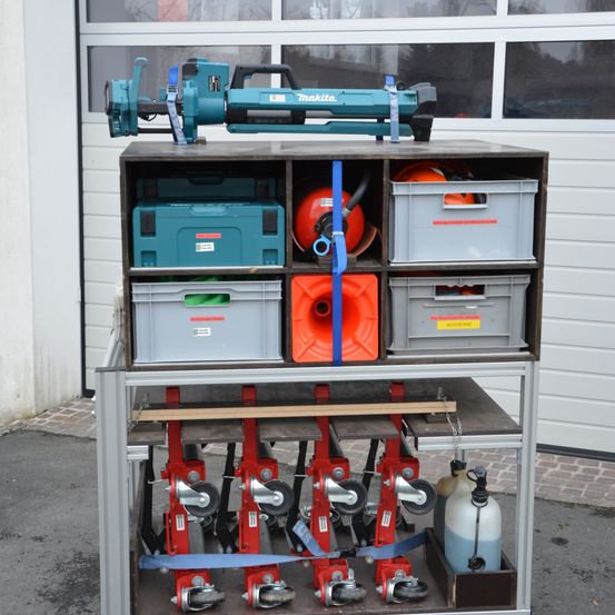 Ein Makita-Werkzeug, das auf einem mehrstöckigen Metallwagen mit verschiedenen Lagerboxen und einem roten Feuerlöscher montiert ist. Unter dem Werkzeug befinden sich rote Radheber, Lagerboxen und ein blauer Tank. Der Wagen steht vor einer Garagentür.