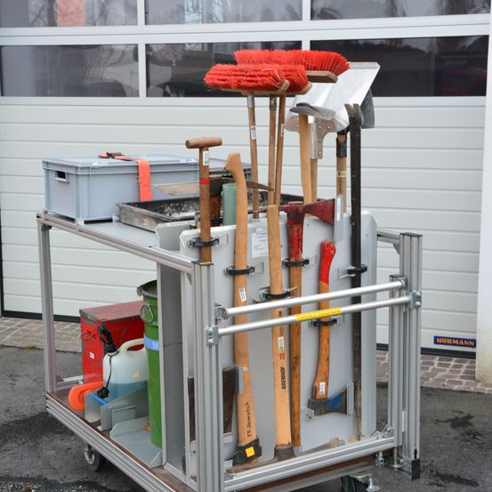 Ein Metallwagen gefüllt mit verschiedenen Werkzeugen, darunter Äxte, Besen und Schaufeln. Der Wagen steht vor einer Garage.