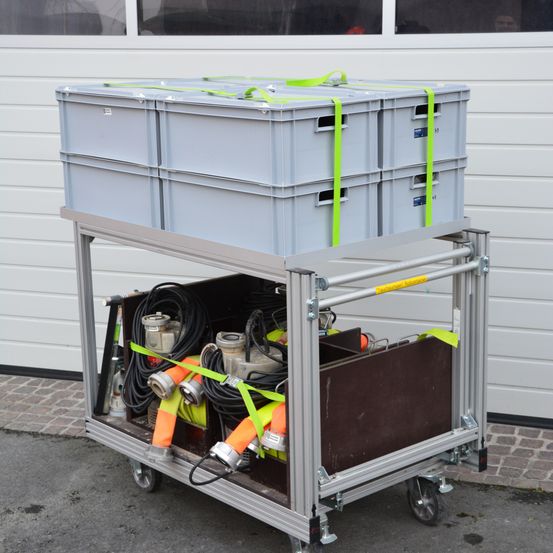 Ein grauer Trolley mit mehreren Aufbewahrungsboxen und Ausrüstung, mit grünen Gurten gesichert, vor einer Garage platziert.