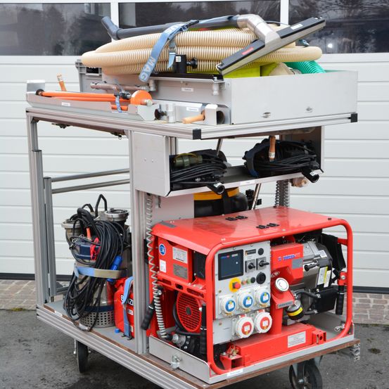 Ein roter und grauer Maschinenwagen mit verschiedenen Werkzeugen und Schläuchen steht vor einem Gebäude. Der Wagen hat mehrere Ebenen, auf denen Ausrüstung verstaut ist.