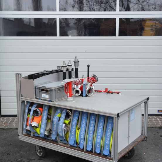 Ein Metallwagen mit Feuerwehrausrüstung, darunter Rohre und Ventile, steht vor einer Garage. Der Wagen hat eine graue Oberseite und einen Metallrahmen.