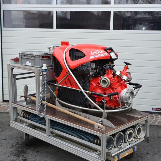 Ein roter Feuerwehrwagen mit dem Wort Magirus darauf, steht vor einer Garage. Es gibt Werkzeuge und Rohre auf dem Wagen.