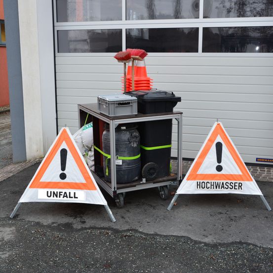 Zwei orangefarbene und weiße Dreiecksschilder liegen auf dem Boden. Eines sagt 'UNFALL' und das andere 'HOCHWASSER'. Hinter den Schildern steht ein Wagen mit Fässern, Abfalleimern und Reinigungsutensilien. Im Hintergrund befindet sich eine Garagentür.