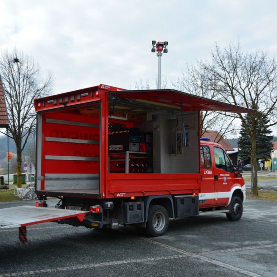 Ein roter Nutzfahrzeug mit offenem Heck und einer Rampe steht am Straßenrand. Im Hintergrund befinden sich Bäume und ein Gebäude.