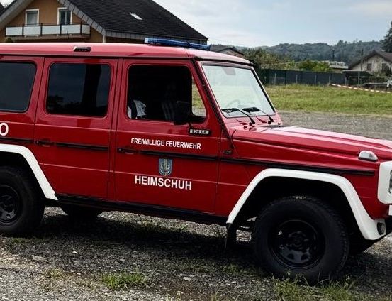 Ein rotes Fahrzeug mit 'KDO' und 'FREIMILLIGE FEUERWEHR' Texten auf der Seite ist in einem Kiesbereich in der Nähe von Häusern geparkt.