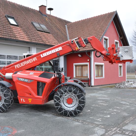Ein roter Liebherr-Traktor mit Ketten an den Reifen steht vor einem roten Haus mit braunem Dach und mehreren Fenstern.