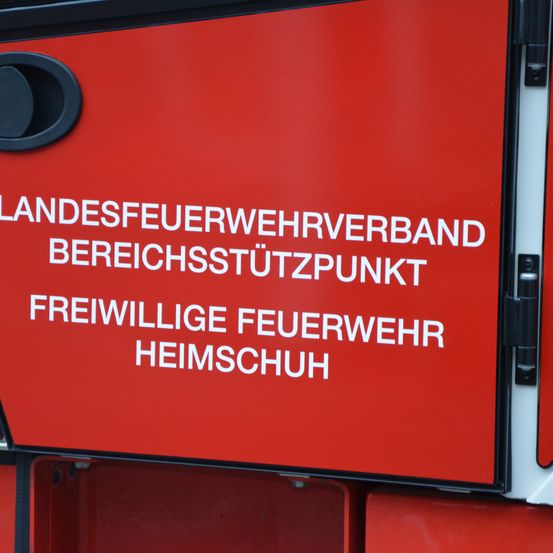 Ein rotes Schild mit weißem Text zeigt 'LANDESFEUERWEHRVERBAND BEREICHSSSTUTZPUNKT FREIWILLIGE FEUERWEHR HEIMSCHUH'. Das Schild ist an einem schwarzen Objekt befestigt.