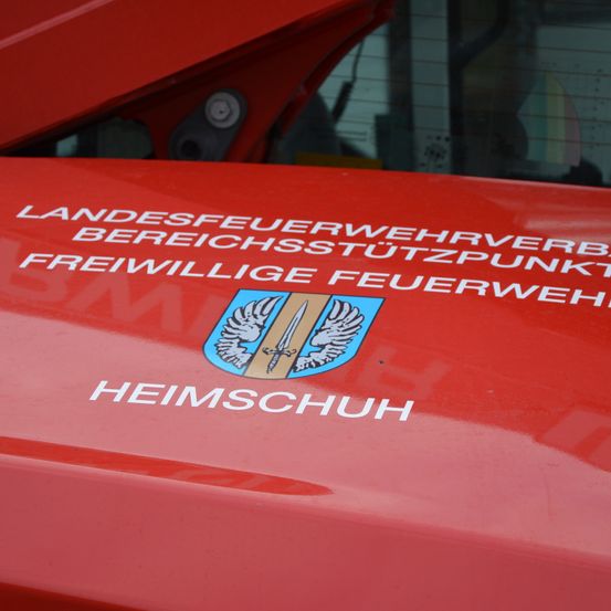 Ein rotes Fahrzeug zeigt den Text 'LANDESFEUERWEHRVERBAND BEREICHSTÜTZPUNKT FREIWILLIGE FEUERWEH HEIMSCHUH'. Unten befindet sich ein Logo mit Flügeln und einem Schwert. Der Schriftzug 'Heimschuh' steht in weiß auf der roten Fläche.