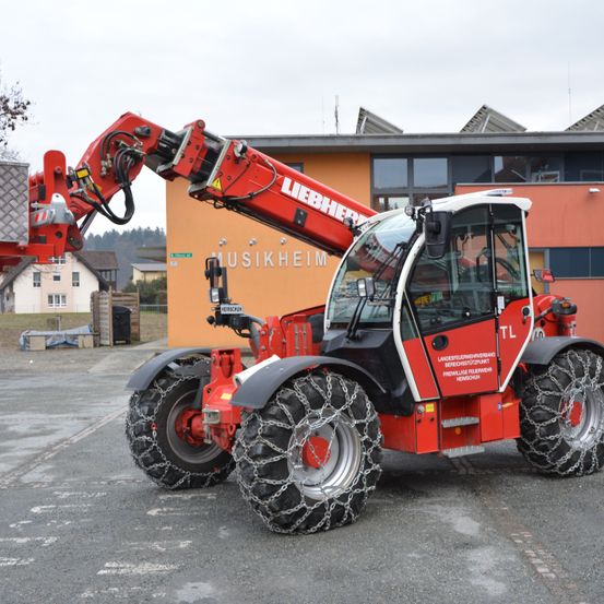 Ein roter Liebherr-Teleskoplader mit Ketten an den Reifen steht vor einem Gebäude mit Glasfenstern. Das Gebäude trägt die Aufschrift 'MUSIKHEIM'.