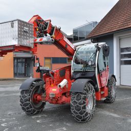 Ein roter Liebherr-Teleskoplader mit einer silbernen Werkzeugkiste steht vor einem Haus. Das Fahrzeug hat Schneeketten an den Reifen und ist mit einem hydraulischen Kran ausgestattet.