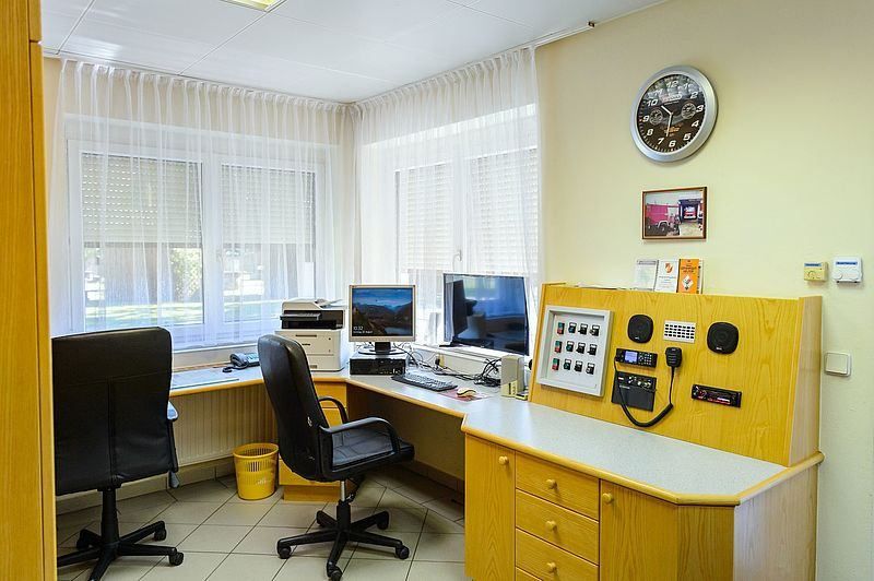 Ein Büroraum mit zwei Computermonitoren, einem Drucker und einer Uhr an der Wand. Es gibt zwei Stühle und einen Schreibtisch mit einem Telefon.