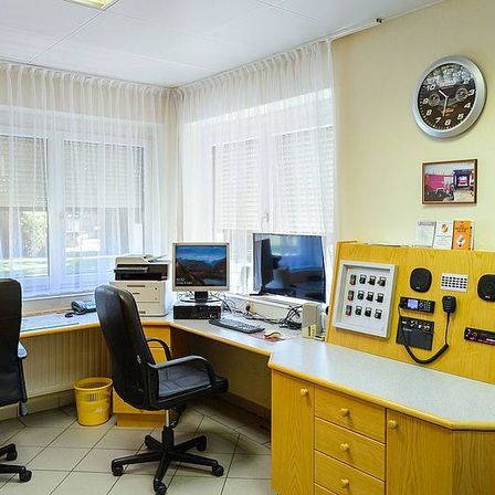 Ein Büroraum mit zwei Computermonitoren, einem Drucker und einer Uhr an der Wand. Es gibt zwei Stühle und einen Schreibtisch mit einem Telefon.