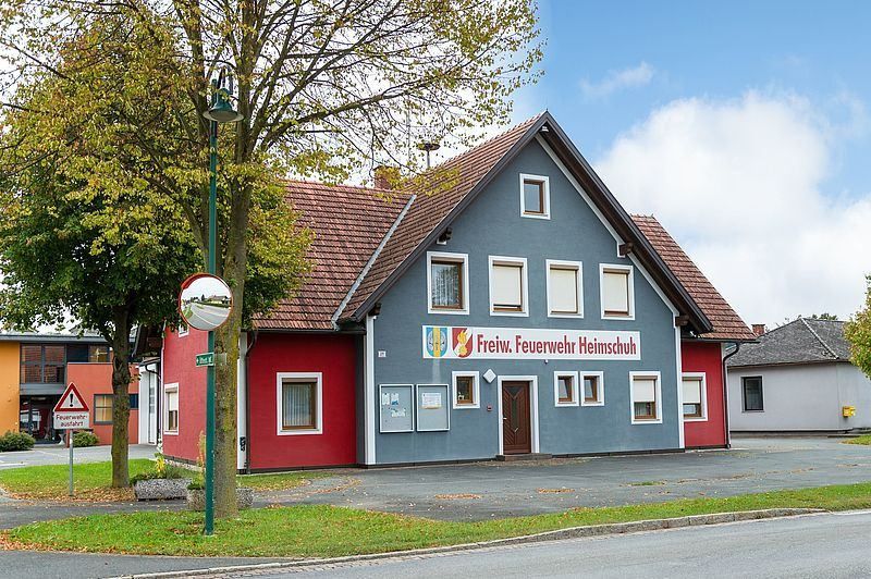 Ein rot-blaues Gebäude mit der Aufschrift Freiw. Feuerwehr Heimschuh. Es hat ein braunes Dach und einen Schornstein. Bäume, eine Straßenlaterne und ein Schild sind in der Gegend. Der Himmel ist klar mit Wolken.