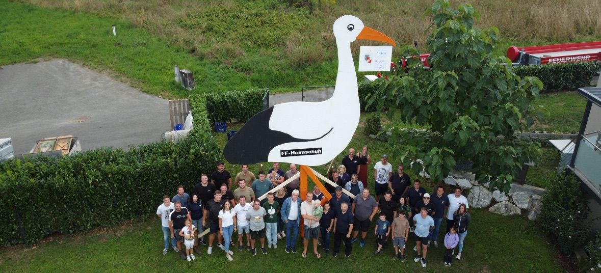 Eine Gruppe von Menschen steht vor einer großen Storchstatue in einem Park. Der Storch hat einen Text, der 'FF-Heimschuh' lautet.