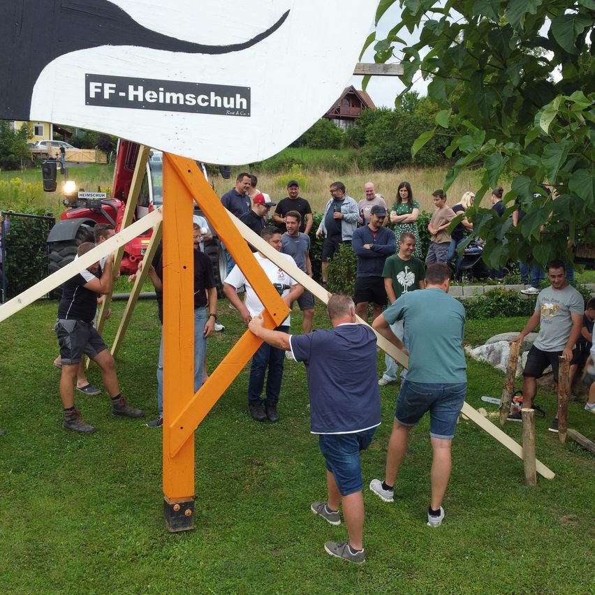 Mehrere Personen arbeiten gemeinsam an einer Holzstruktur in einem Garten, während Zuschauer zuschauen. Die Struktur hat ein Schild mit der Aufschrift FF-Heimschuh.