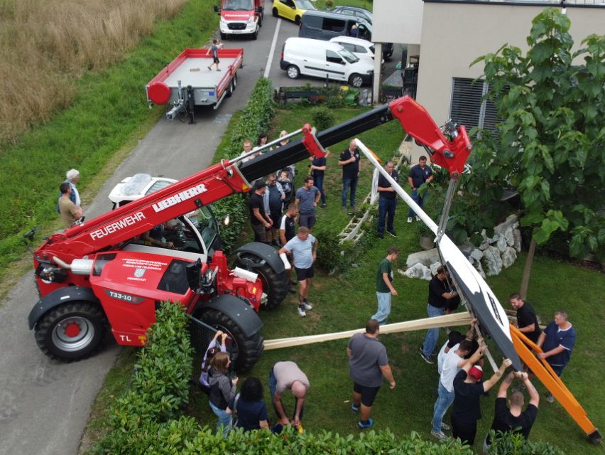 Eine Gruppe von Menschen benutzt einen roten Kran, um ein großes weißes und schwarzes Objekt, möglicherweise ein Boot, in die Luft zu heben. Sie sind von Grünpflanzen und geparkten Fahrzeugen umgeben, einschließlich eines weißen Vans.
