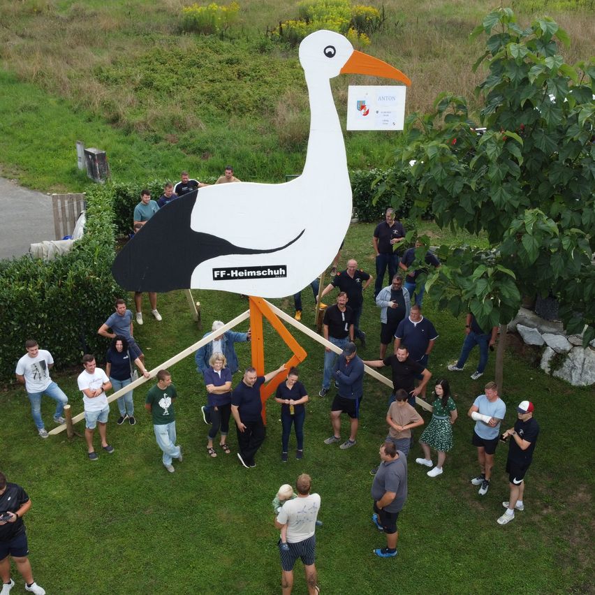 Eine Gruppe von Menschen versammelt sich um eine große Storch-Skulptur. Einige halten Holzbretter in der Hand, möglicherweise beim Zusammenbau. Die Szene spielt im Freien mit Rasen und einem Schild hinter dem Storch.