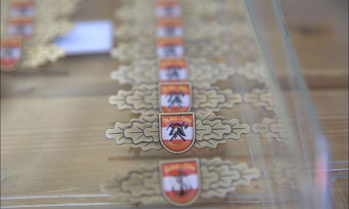 Eine Reihe von goldfarbenen Emblemen mit komplizierten Blattdesigns, jedes mit einem rot-weißen Schild. Das zentrale Emblem zeigt einen Vogel und die Worte 'OLRG-OBV'. Der Hintergrund ist unscharf.