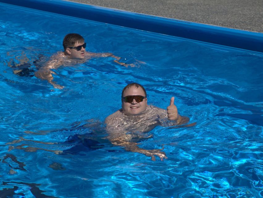 Zwei Männer schwimmen in einem blauen Pool, einer gibt einen Daumen hoch. Beide tragen Sonnenbrillen.
