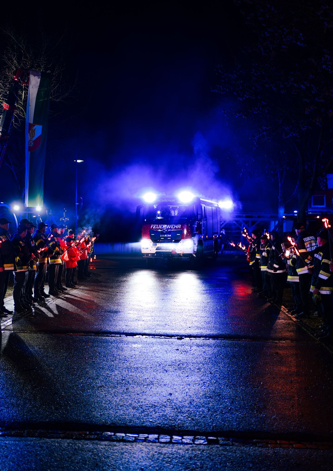 Ein Feuerwehrauto fährt mit blauen Lichtern und Rauch auf der Straße. Es stehen Feuerwehrleute auf beiden Seiten der Straße.