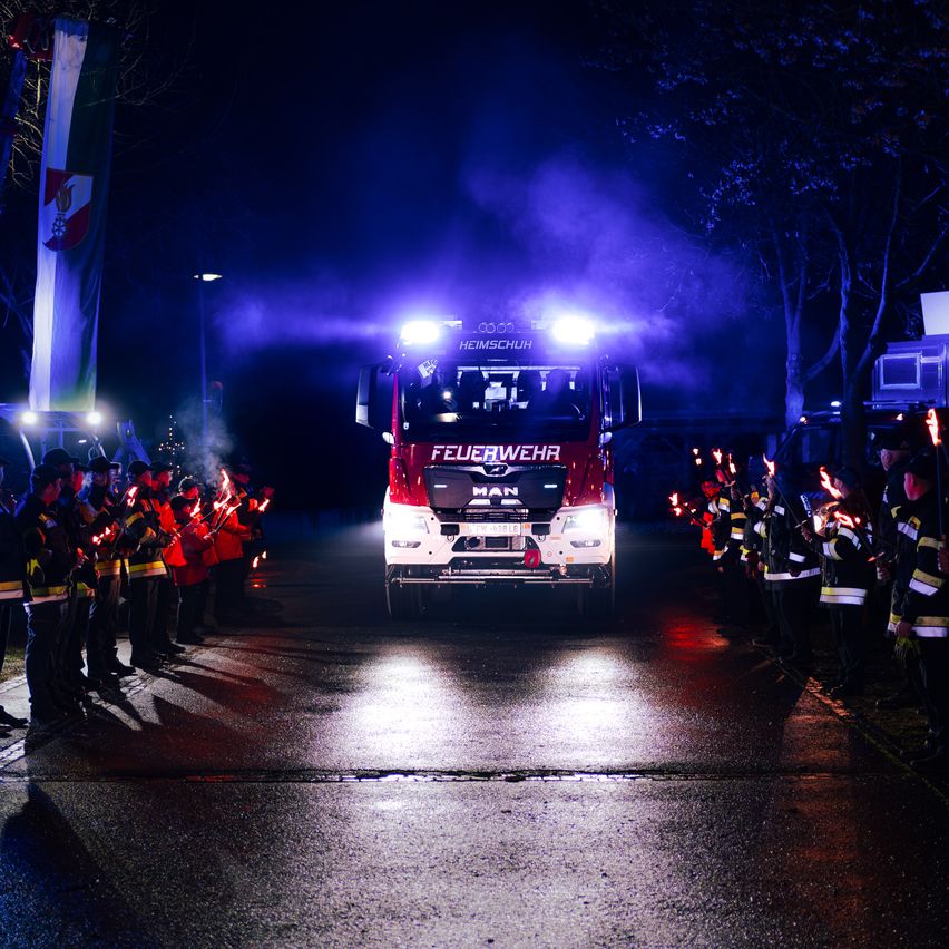 Ein Feuerwehrauto fährt nachts auf einer Straße, wobei Feuerwehrleute auf beiden Seiten Fackeln halten und Rauch aus dem Fahrzeug aufsteigt.