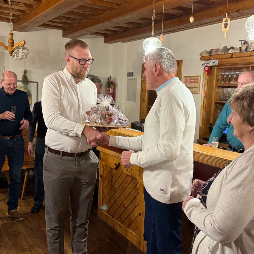 Zwei Männer tauschen ein Geschenk in einem Raum aus. Der Mann auf der linken Seite lächelt und hält ein verpacktes Geschenk. Der Mann auf der rechten Seite schaut auf das Geschenk. Eine Frau steht neben dem Mann auf der rechten Seite. Im Hintergrund stehen Personen in der Nähe einer Theke mit Weingläsern und Flaschen.