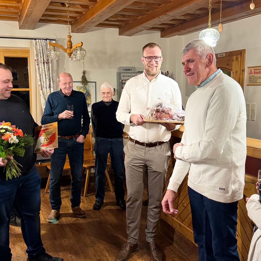 Eine Gruppe von Männern versammelt sich drinnen und lächelt. Ein Mann präsentiert ein eingewickeltes Geschenk. Ein anderer hält einen Blumenstrauß. Ein Schild ist im Hintergrund sichtbar.