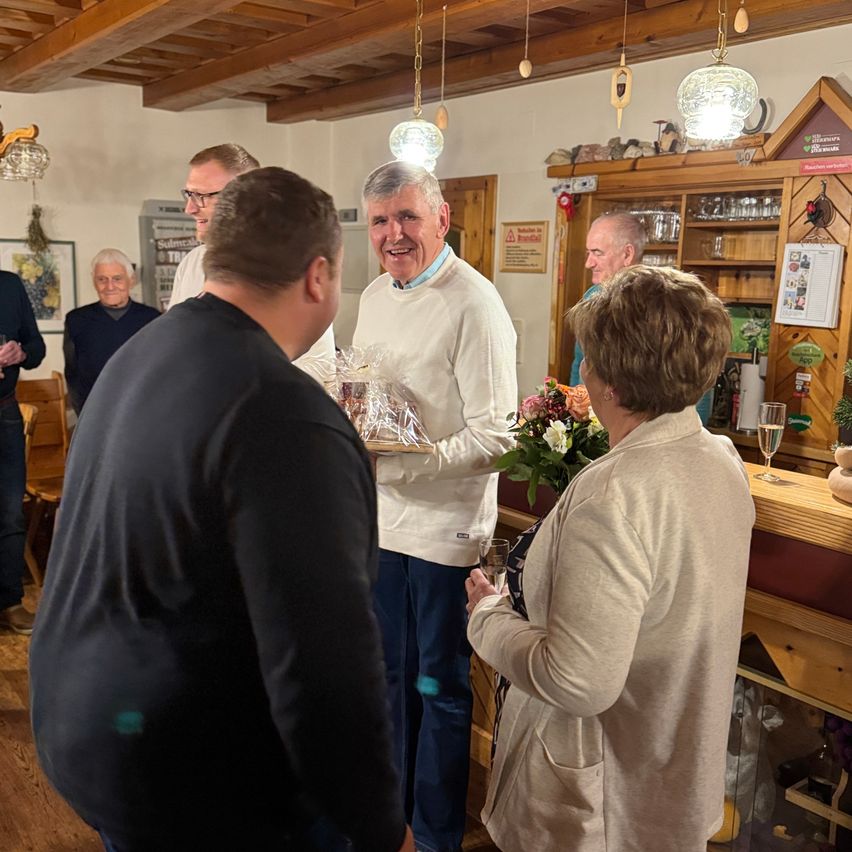 Mehrere Personen stehen in einem Raum, ein Mann in Weiß präsentiert einem Mann in Schwarz ein verpacktes Geschenk. Eine Frau mit kurzen Haaren hält ein Weinglas. Hinter ihnen schaut eine Frau mit Blumen zu.