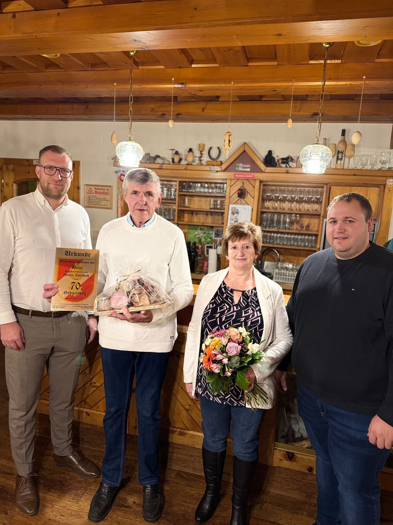 Vier Personen stehen in einem Raum. Zwei Männer sind auf beiden Seiten, während eine Frau in der Mitte Blumen hält. Der Mann auf der linken Seite hält ein Zertifikat und der Mann auf der rechten Seite einen Korb mit Essen.