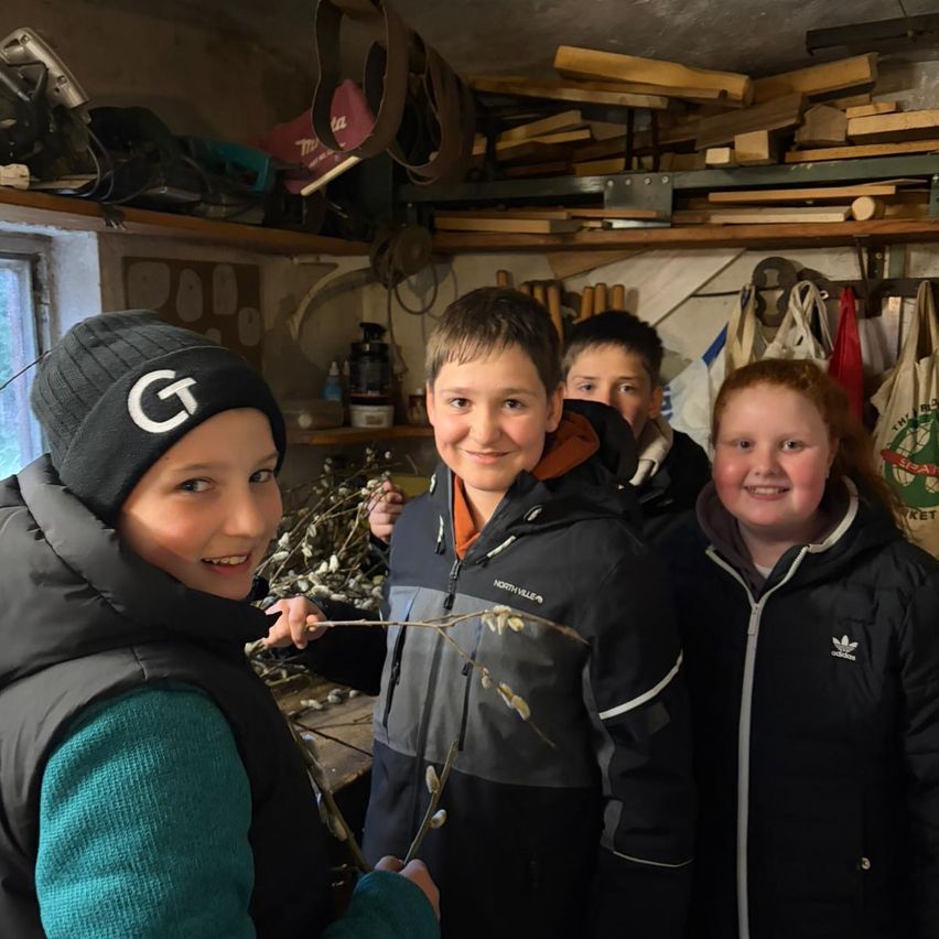 Vier Kinder in Winterjacken stehen in einem Raum, eines hält einen Zweig mit weißen Blüten, zwei Regale mit verschiedenen Gegenständen im Hintergrund.