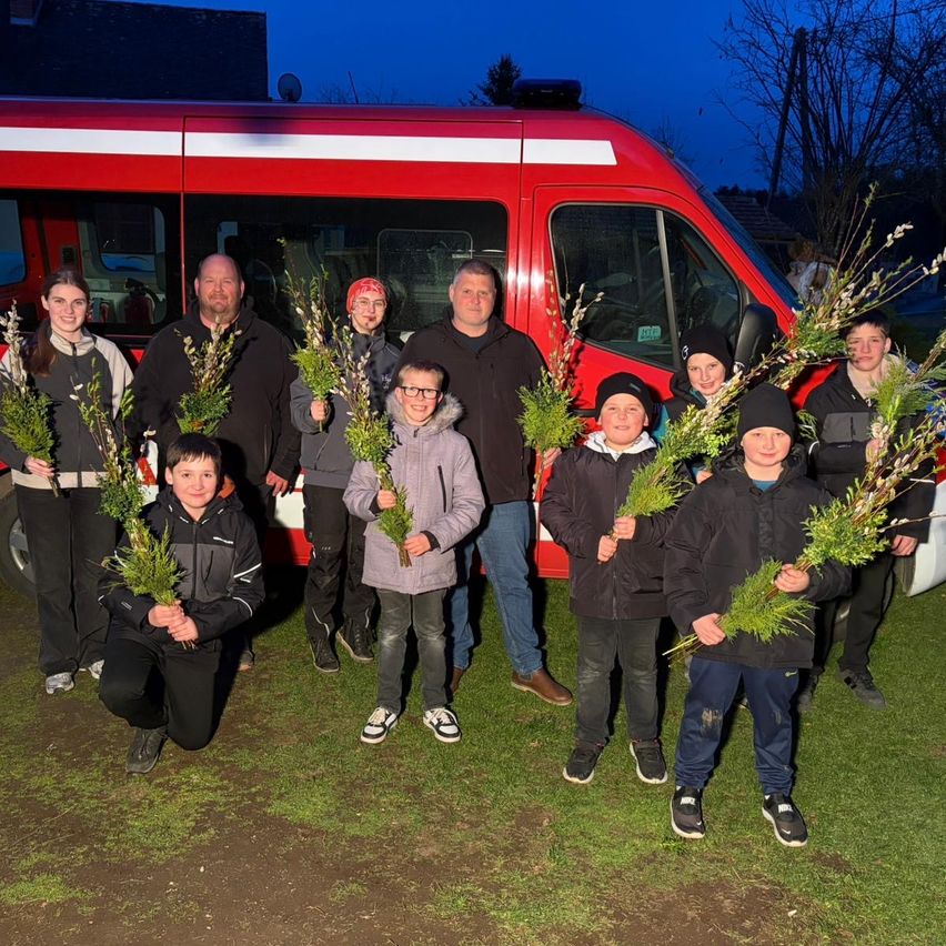 Eine Gruppe von Menschen steht draußen mit einem roten Van hinter ihnen, hält grüne Zweige, lächelt und posiert für ein Foto in der Dämmerung.