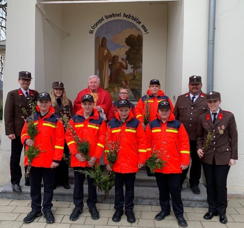Eine Gruppe von Menschen in Uniformen mit Hüten posiert für ein Foto vor einem Gebäude. Sie halten Blumen. Dahinter ist ein Gemälde einer religiösen Szene.