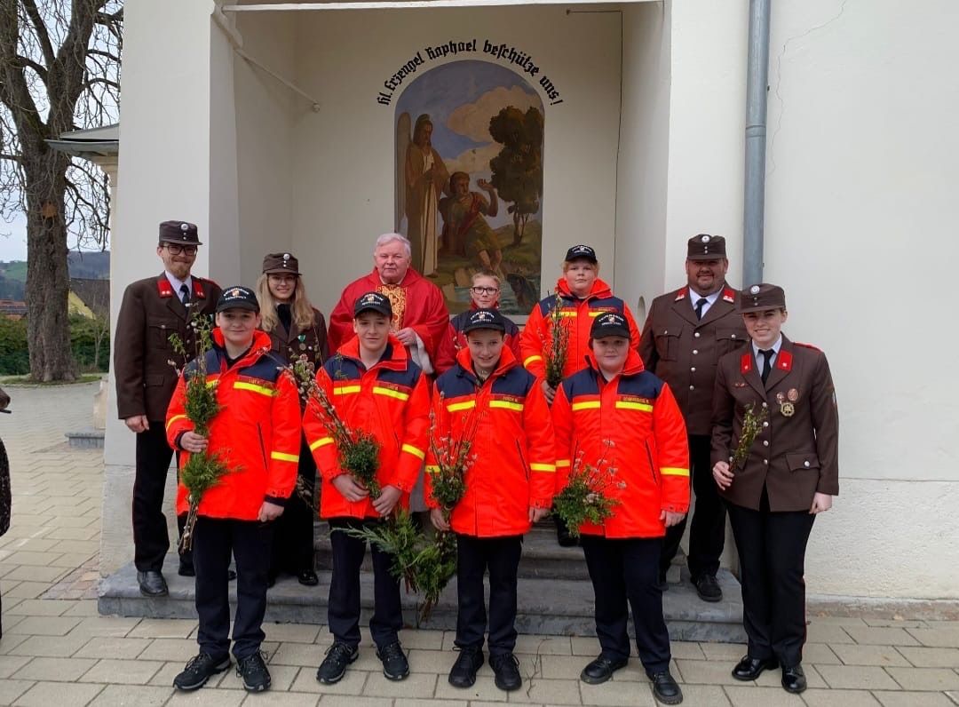 Eine Gruppe von Menschen in Uniformen mit Hüten posiert für ein Foto vor einem Gebäude. Sie halten Blumen. Dahinter ist ein Gemälde einer religiösen Szene.