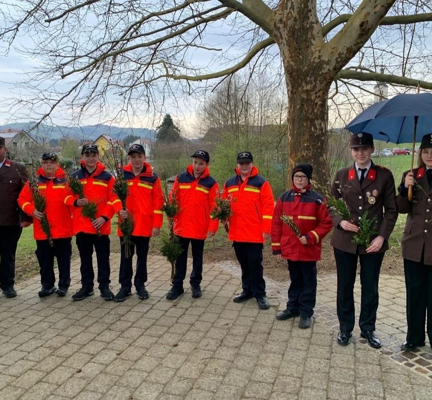 Eine Gruppe Feuerwehrleute, gekleidet in roten und gelben Uniformen, stehen zusammen und halten Pflanzen. Sie stehen unter einem Baum auf einem gepflasterten Bereich mit einem Gebäude und Bergen im Hintergrund.